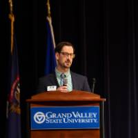 Man in suit gives speech on stage behind podium, Groundswell staff in background